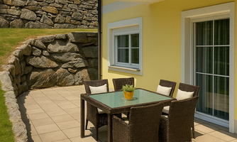 A terrace with a table and chairs made of rattan. In the background, you can see a green landscape and a yellow house wall.