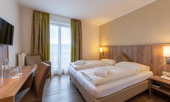 A modern hotel room with two beds and a desk. Large windows let in plenty of daylight.