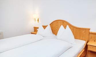 A cozy room with a wooden double bed and white bedding. Next to the bed, there is a bedside lamp and wooden shelves.