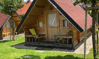 A cozy wooden house with a red gabled roof. It stands on a green meadow, surrounded by small trees and outdoor seating areas.
