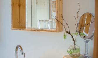 A modern bathroom with a round glass sink and a wooden mirror. On the shelf, there are care products and a small branch with fresh leaves.