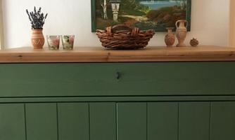 A green cupboard with a wooden top. Above it hangs a painting of mountains and a lake, and there are some pots and a bowl on it.