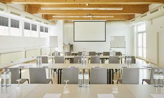 A modern conference room with tables and chairs.  
On the wall, there is a large screen, and there are water glasses and bottles on the tables.