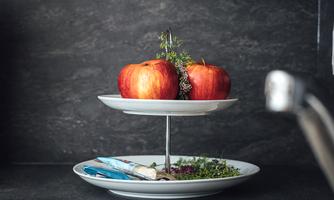 A stylish kitchen area with a two-tier serving stand. The upper plate holds red apples, while the lower plate is decorated with herbs.