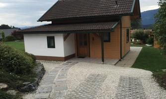 A small wooden house with a gable roof and a terrace. The paved path leads to the entrance and is surrounded by green plants.