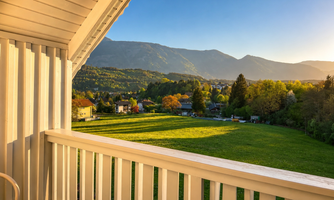 A balcony view of a green meadow and gentle hills in the background. The scene is illuminated by warm sunlight and a clear sky.
