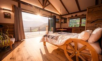 A cozy room with wooden furniture and a large window that offers a beautiful view of the mountains. The sun is shining through the window, creating a warm atmosphere.