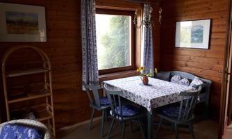 A cozy dining area with a wooden table, surrounded by chairs. Daylight enters through the window, and the walls are clad in wood.