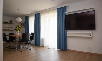 A bright living room with large windows and blue curtains. In the corner, there is a dining table with chairs and a television on the wall.