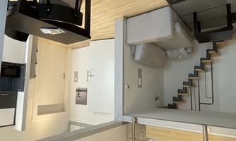 A modern, bright apartment with a living room, a small kitchen, and a staircase to an upper floor. The space is minimally furnished and feels inviting.