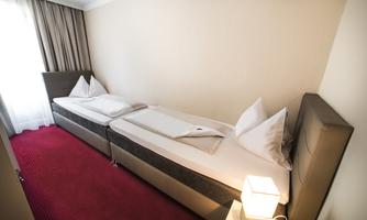 A simple hotel room with two single beds and a red carpet. The room has a window and a modern bedside lamp.