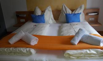 A cozy bed with white pillows and blue decorative cushions. On the bedspread, there is an orange cloth and folded towels.