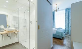 A modern bathroom with a glass shower enclosure and a sink. In the background, a bright bedroom is visible with a bed and blue curtains.