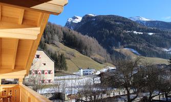 A mountain landscape with snow-covered peaks and green meadows. In the foreground, a wooden balcony is visible.