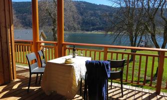 A terrace with a view of a lake and hills. A table with a tablecloth, two chairs, and a hat is set.