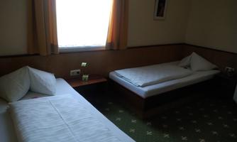 A simple room with two single beds and a window through which daylight streams. The floor is carpeted in green, and there is a bedside lamp.