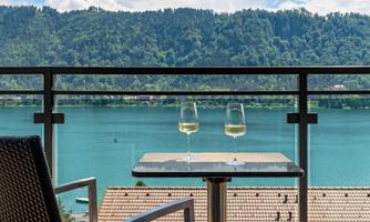 A beautiful view of a clear lake with mountains in the background. On the balcony, there are two wine glasses on a table.