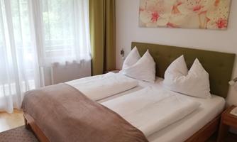 A bright, cozy bedroom with a large bed and two pillows. The room features large windows with curtains and a floral decoration on the wall.