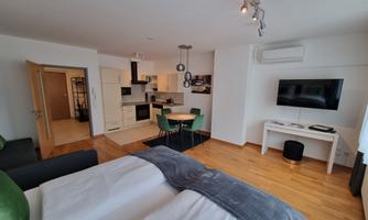 A modern room with a double bed, a kitchenette, and a dining area. The space is bright and friendly, with wooden flooring and large windows.