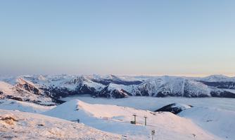 An impressive mountain landscape with snow-covered peaks and a clear sky. In the foreground, there are snow mounds and ski lifts.