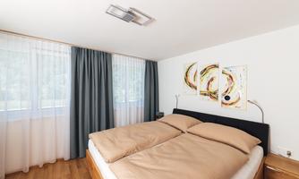 A modern bedroom with a large double bed and soft beige tones. The windows are adorned with bright curtains, and artistic pictures hang on the wall.