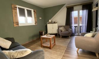 A cozy living room with two comfortable sofas, a wooden table, and a carpet. The walls are painted green, and there are large windows overlooking the outside.