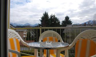 A cozy balcony with two chairs and a round table. In the background, mountains and a cloudy sky are visible.