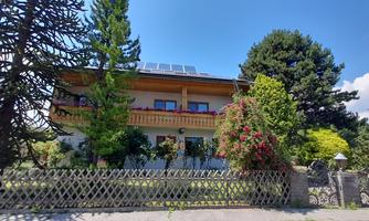 A beautiful house with a balcony and solar panels on the roof. The garden is lush and green, surrounded by trees and flowers.