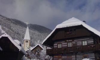 A snowy village in the mountains with traditional wooden chalets. In the background, a church tower is visible, surrounded by snow-covered trees.
