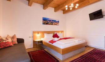 A cozy hotel room with a wooden beam ceiling, a double bed, and a sofa. A picture of a beautiful landscape hangs on the wall.