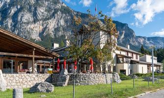 A cozy building in the Alps with a stone facade and large windows. In the background, majestic mountains stretch out under a clear sky.