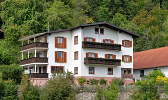 A typical residential house in a rural setting with several balconies. The facade is white, surrounded by green foliage and trees.