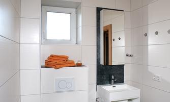 A modern bathroom with white tiles and a large mirror. On the shelf, there are orange towels and there is a sink as well as a toilet.