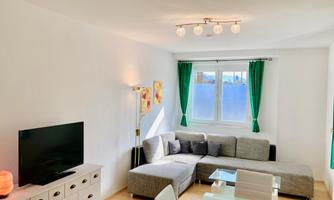 A bright and modern living room with a gray couch and a television. Large windows with green curtains allow for plenty of light.