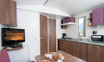 A modern kitchen with wooden cabinets, a table, and a television. On the table are drinks and pastries.