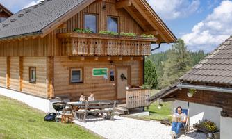 A charming wooden house with a balcony and blooming plants. People are sitting at a table in front of the house, enjoying the outdoor atmosphere.