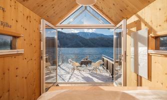 A modern room with wood interior and a large window that offers a view of a lake and snow-capped mountains. Outside, two chairs and a table are placed on the terrace.