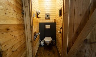 A small, rustic toilet room with paneling. The toilet is simply furnished and has various accessories as well as a water jug.