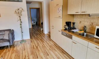 A modern kitchen with wooden flooring and bright furniture. In the background, a cozy living room area is visible.