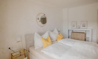 A modern bedroom with a large bed and decorative pillows. A round mirror hangs on the wall, and a wooden shelf stands next to it.