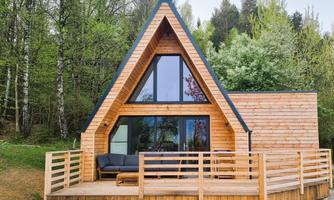 A modern wooden house in A-frame style with large windows. It features a terrace and is surrounded by trees.