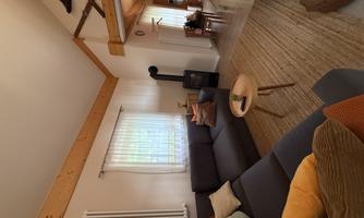 A cozy living room with a gray sofa and a round wooden table. Large windows provide plenty of daylight and a warm atmosphere.