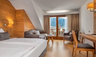 A modern hotel room with wood paneling and large windows. There is a double bed, a cozy seating area, and a desk.