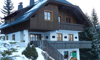 A charming wooden house in the snow with a balcony and green trees in the background. The cozy architecture conveys an inviting atmosphere.