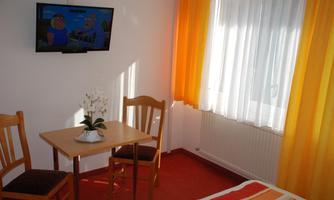 A cozy room with a table, two chairs, and a television on the wall. Windows with light curtains let in a lot of light.