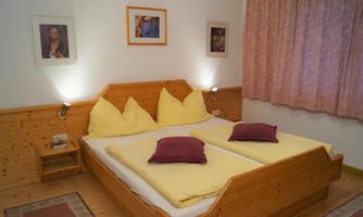 A cozy bedroom with a wooden bed and yellow pillows. The walls are decorated with pictures and the room has curtains in soft colors.