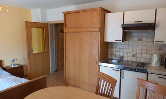 A small, bright apartment with a wooden kitchen and a dining table. On the left is a bed, and a door leads to another room.