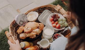 A basket with fresh breakfast, containing croissants, fruit, and drinks. The person is sitting on the ground and enjoying the picnic.