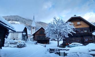 A snow-covered village with traditional wooden houses and a church tower. The surroundings are winterly picturesque and surrounded by snow-covered mountains.