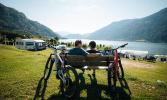 Two people are sitting on a bench and enjoying the view of a lake. In the background, mountains and RVs can be seen.